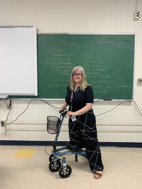 A teacher using a knee scooter in a classroom, standing in front of a chalkboard filled with notes.