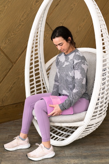 A person wearing a gray and black long-sleeved shirt and purple leggings is sitting in a white wicker hanging chair. They are holding a pink weighted cold pack wrap on their leg. The scene is indoors, with wooden paneling in the background.