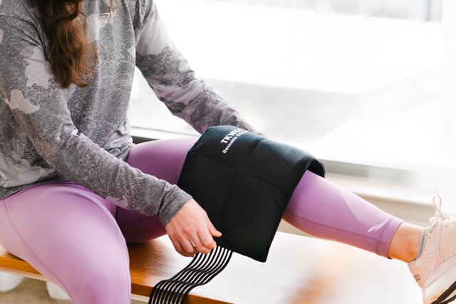 A person wearing a gray and black long-sleeved shirt and purple leggings is sitting on a wooden bench, placing a black weighted cold pack wrap on their leg. The scene is indoors, with a large window in the background.