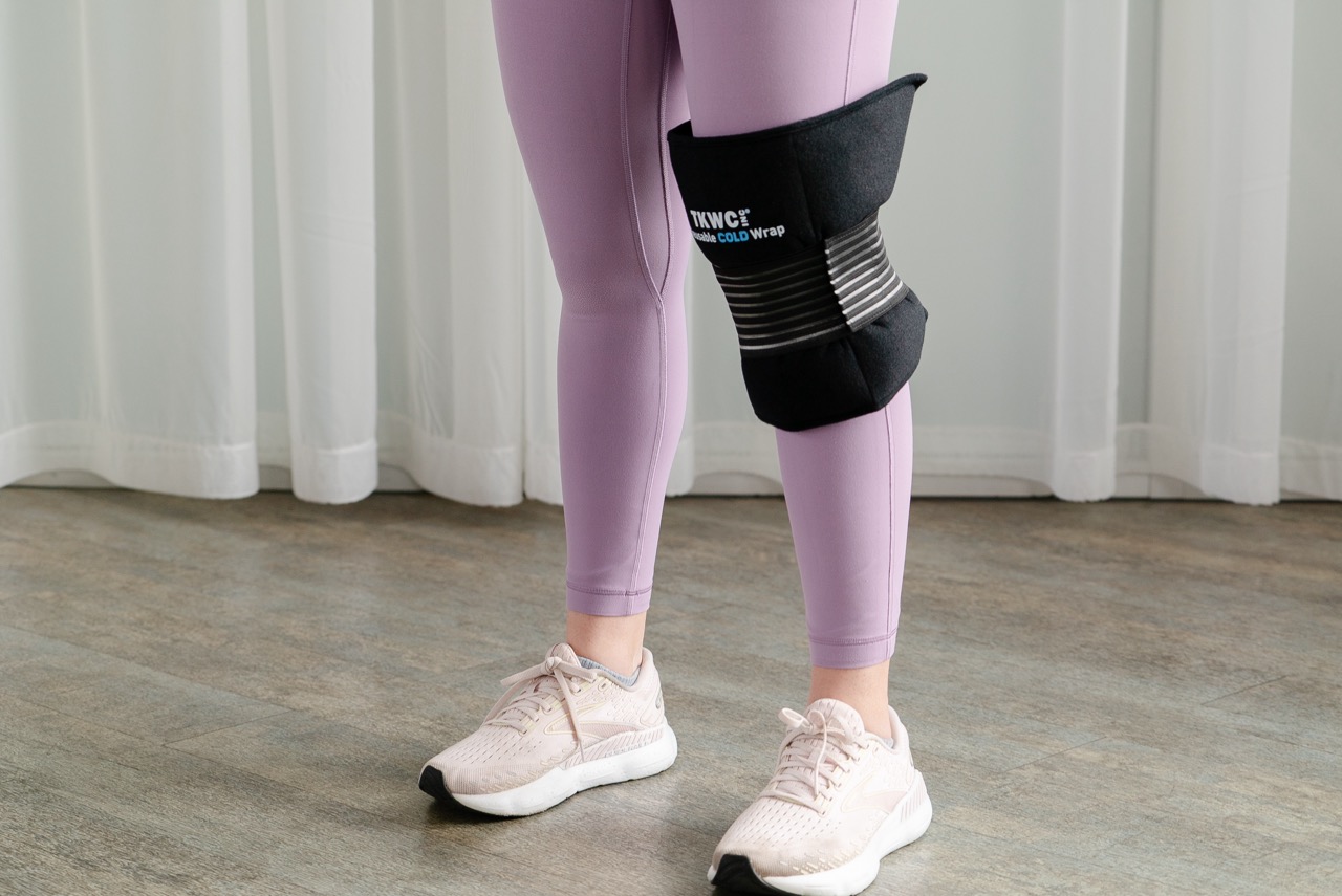 A person standing indoors wearing purple leggings and pink sneakers, with a black weighted cold pack wrap secured around their knee. The background features white curtains.