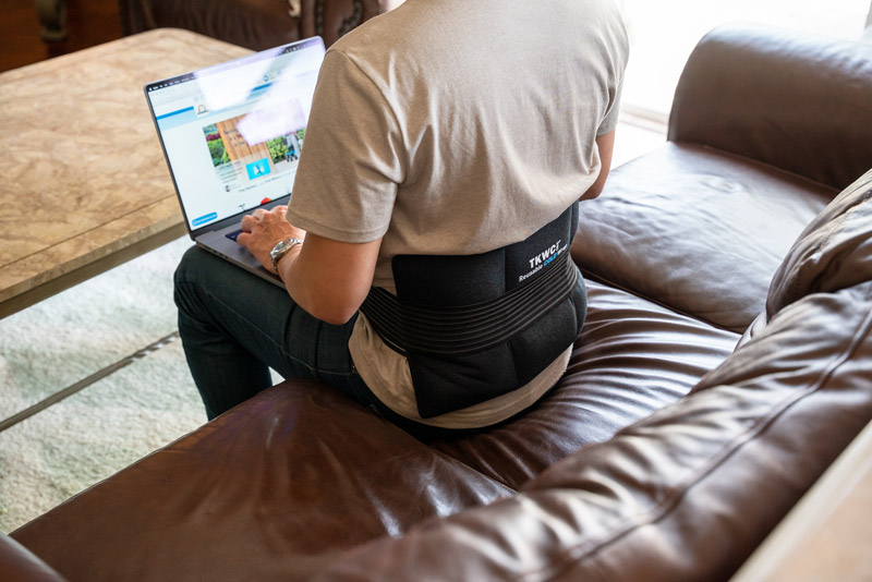 A person sitting on a brown leather couch, working on a laptop. They are wearing a grey shirt and a black back support brace labeled 