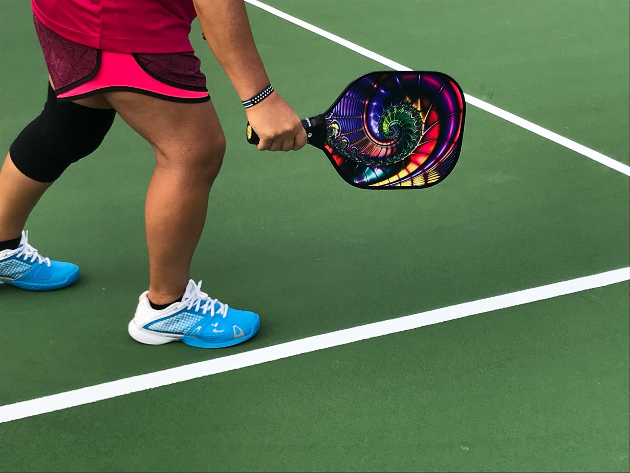 A pickleball player standing on a green outdoor court, wearing bright blue shoes and a knee brace, holding a paddle with a vibrant spiral design.