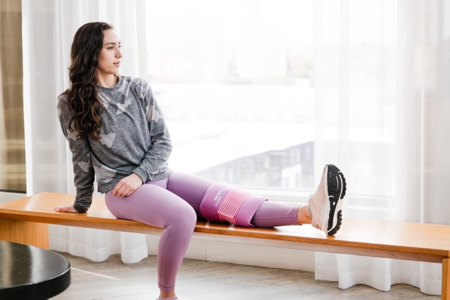 Person sitting on a wooden bench wearing a gray camo-patterned sweatshirt and purple leggings, with a pink reusable cold wrap around their right knee.