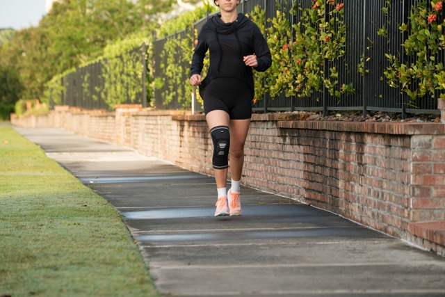 A person wearing a black athletic outfit, black knee brace, white socks, and orange running shoes is jogging on a paved path next to a brick wall and fence with green foliage.