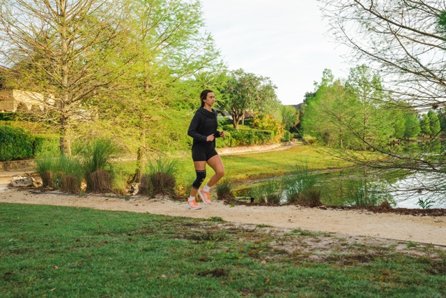 A woman is running along a gravel path by a lake, surrounded by green trees and bushes. She is wearing a black athletic outfit, black knee brace, and pink running shoes.