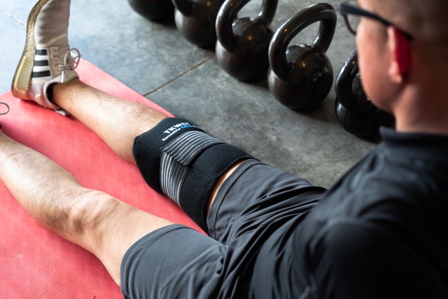 A person is sitting on a red exercise mat, stretching their legs. They are wearing a ice cold pack on the right knee, black shorts, and Adidas sneakers. Kettlebells are lined up in the background.