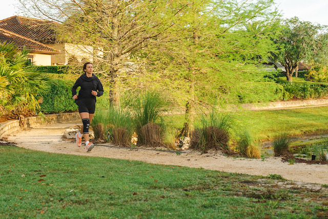 A person jogging on a gravel path in a park-like setting, surrounded by grass, trees, and shrubs, wearing a black knee brace on their right leg.