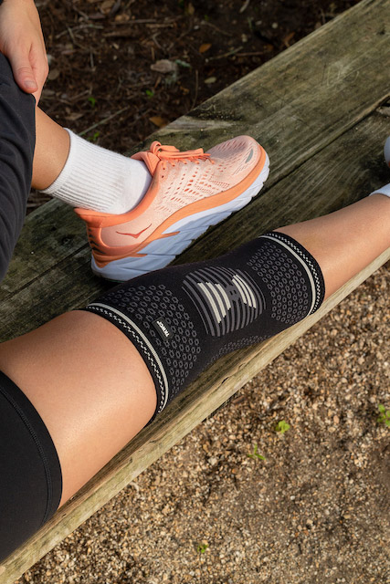 Side view of a black knee compression sleeve  on a person’s right leg, paired with bright orange running shoes on artificial grass.