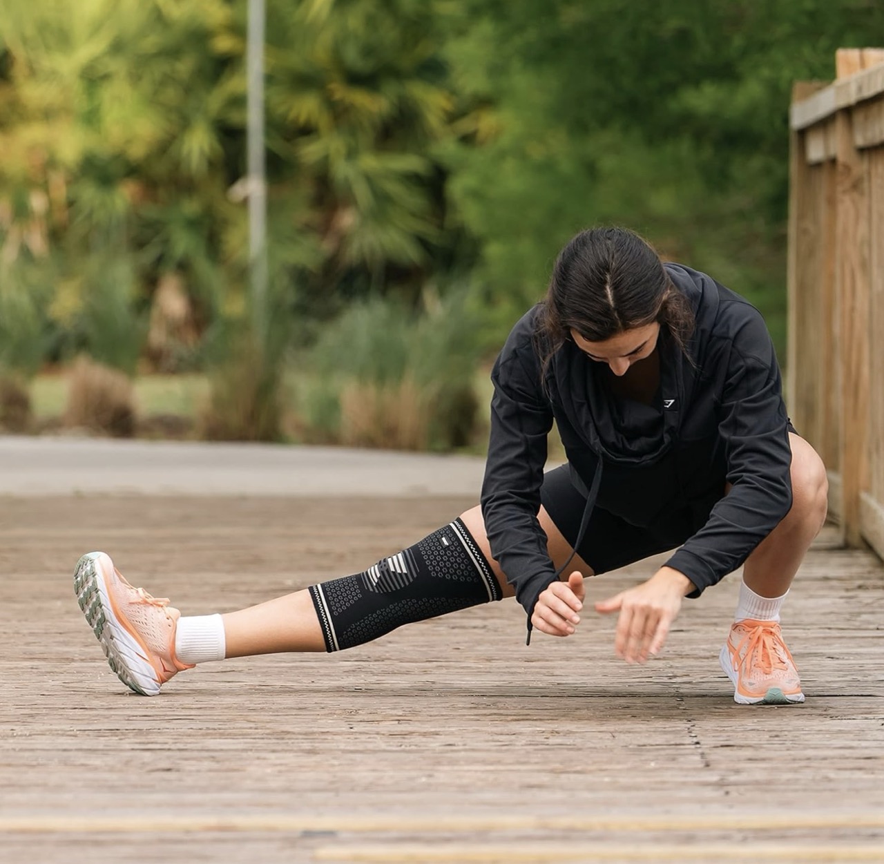 Woman stretching outdoors on a wooden path, wearing a black knee sleeve and athletic wear, surrounded by greenery.