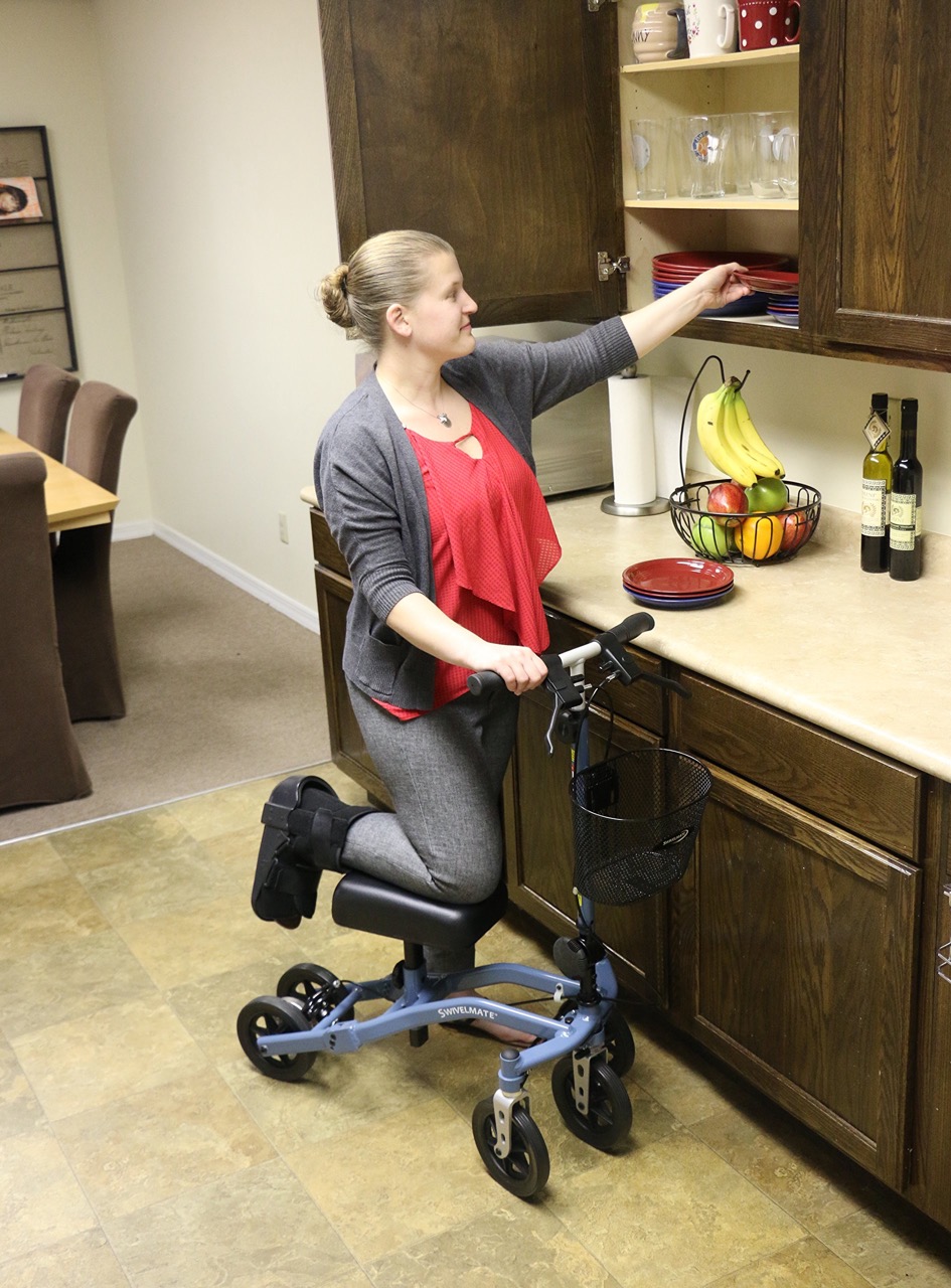 A woman in a kitchen using a knee scooter, with her injured leg elevated on the scooter while she reaches into a cabinet.