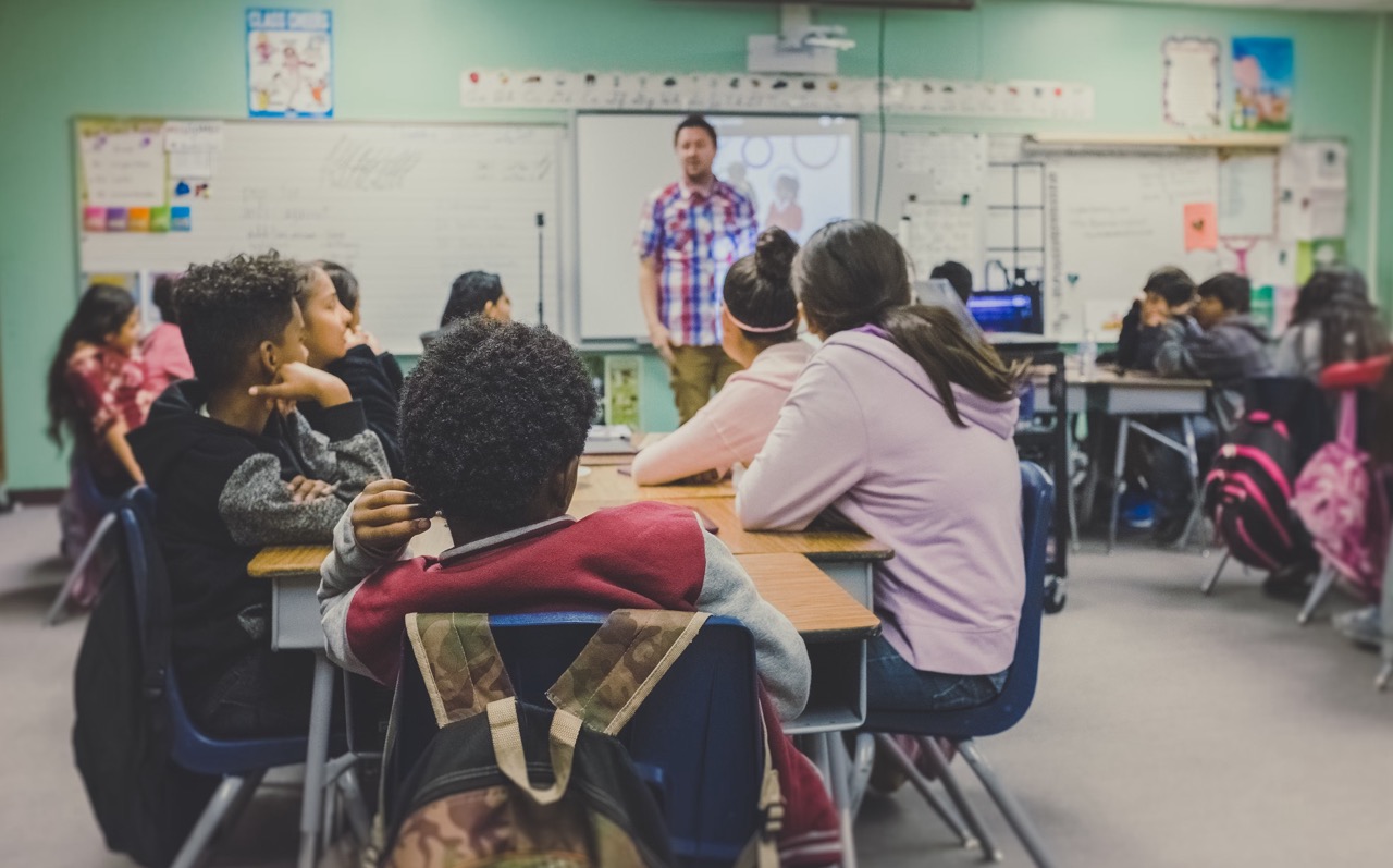 A classroom with students sitting at desks, attentively listening to a teacher who is standing at the front of the room near a whiteboard.