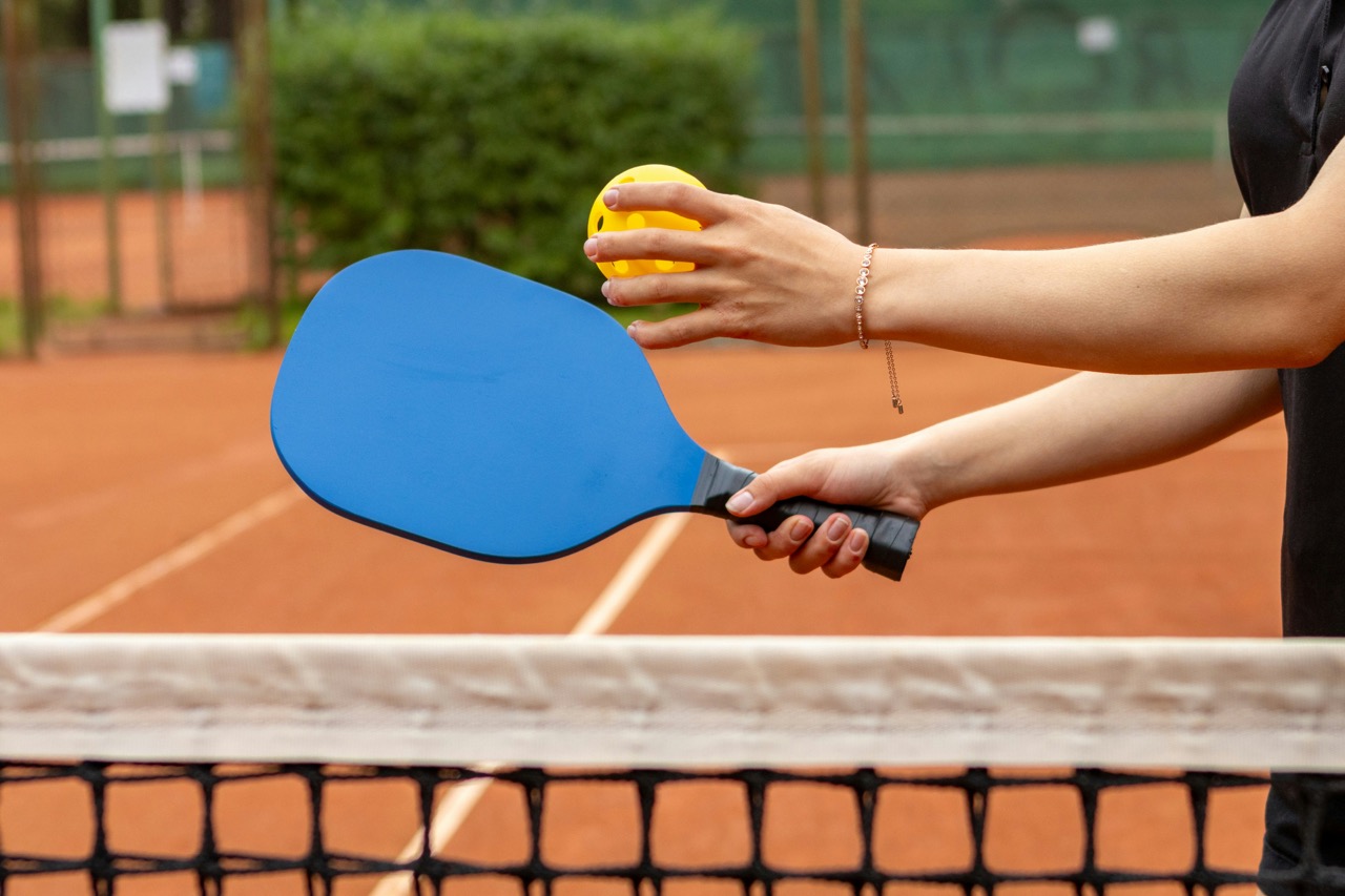 Close-up of a person holding a blue pickleball paddle and a yellow ball, standing by a tennis-style net.