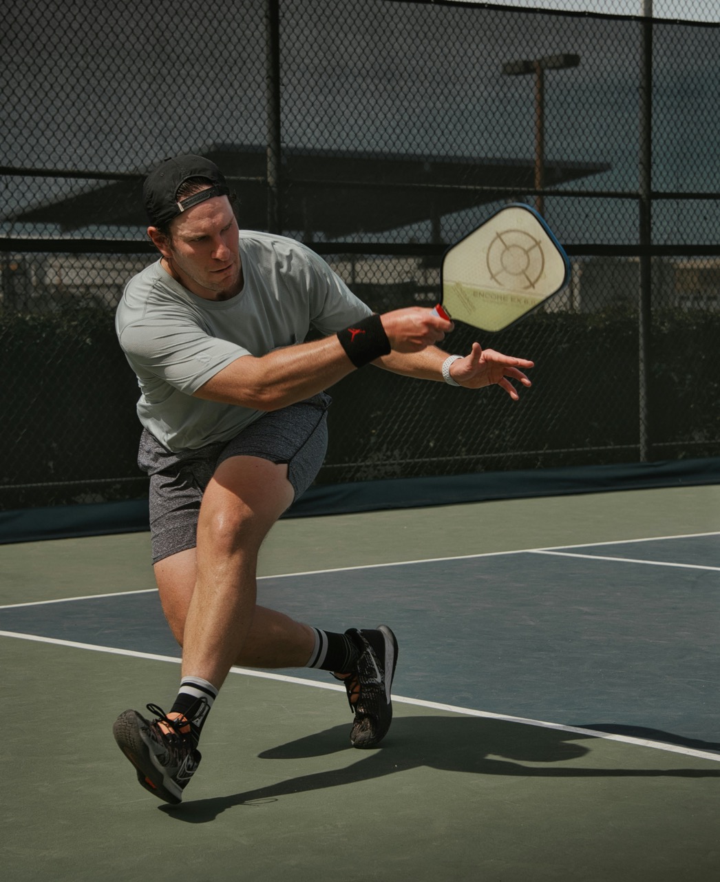 A male pickleball player lunging forward to hit a shot, holding a paddle in his right hand, on an outdoor court.