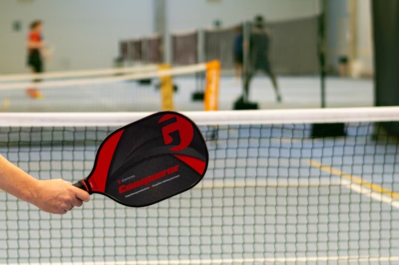 Hand holding a pickleball paddle near the net, with a game in progress in the background.