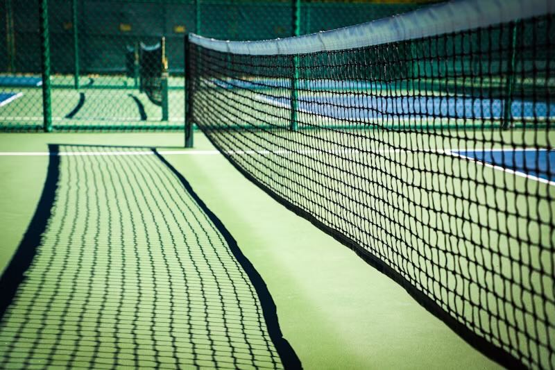 A focused shot of a pickleball net on a court, casting shadows on the green surface. The image highlights the texture and straight alignment of the net, with additional courts visible in the background.