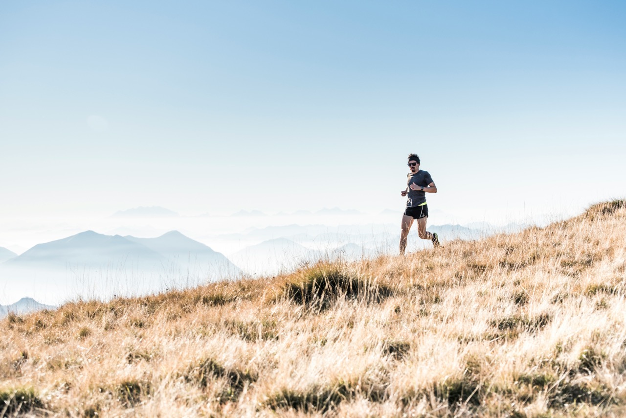 A man running on a grassy hill with distant mountains and a clear blue sky in the background.