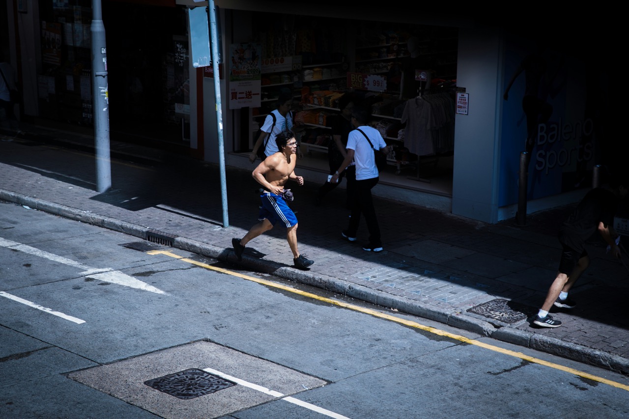 A shirtless man running on a city sidewalk under a shadow, passing by storefronts.