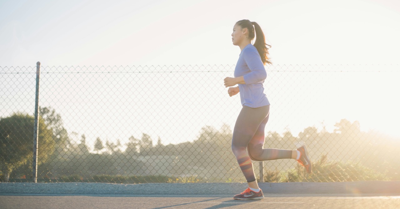 A woman jogging on a paved path next to a chain-link fence, with the sun shining brightly behind her.