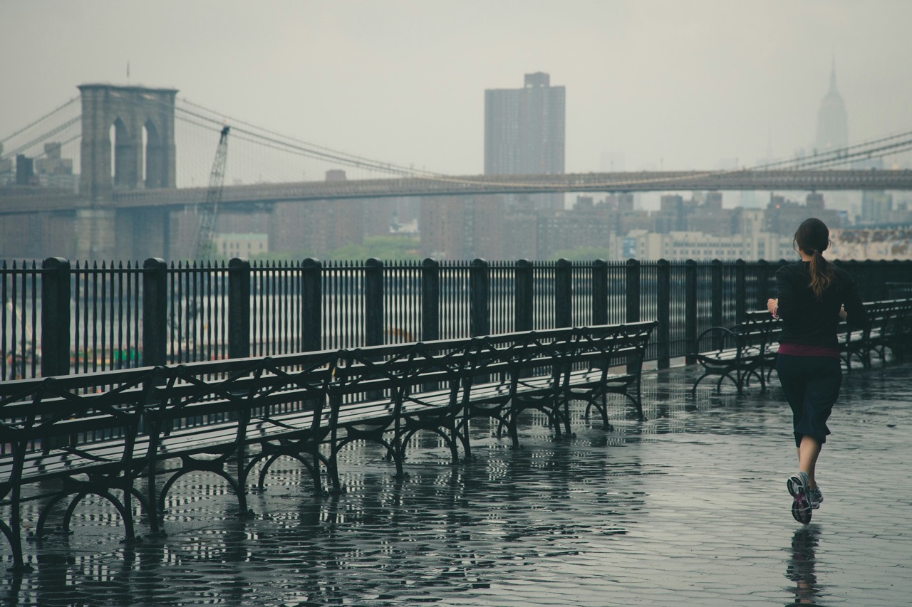 A person jogging along a wet, deserted promenade with the Brooklyn Bridge and city skyline in the background.