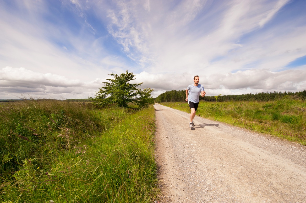 A man running on a gravel road through a green, rural landscape under a partly cloudy sky.