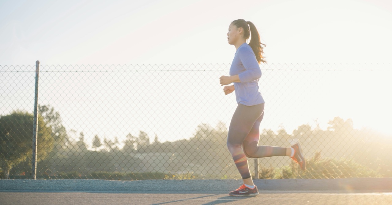 A woman in athletic wear jogs along a paved path near a chain-link fence, with the sun casting a warm glow.