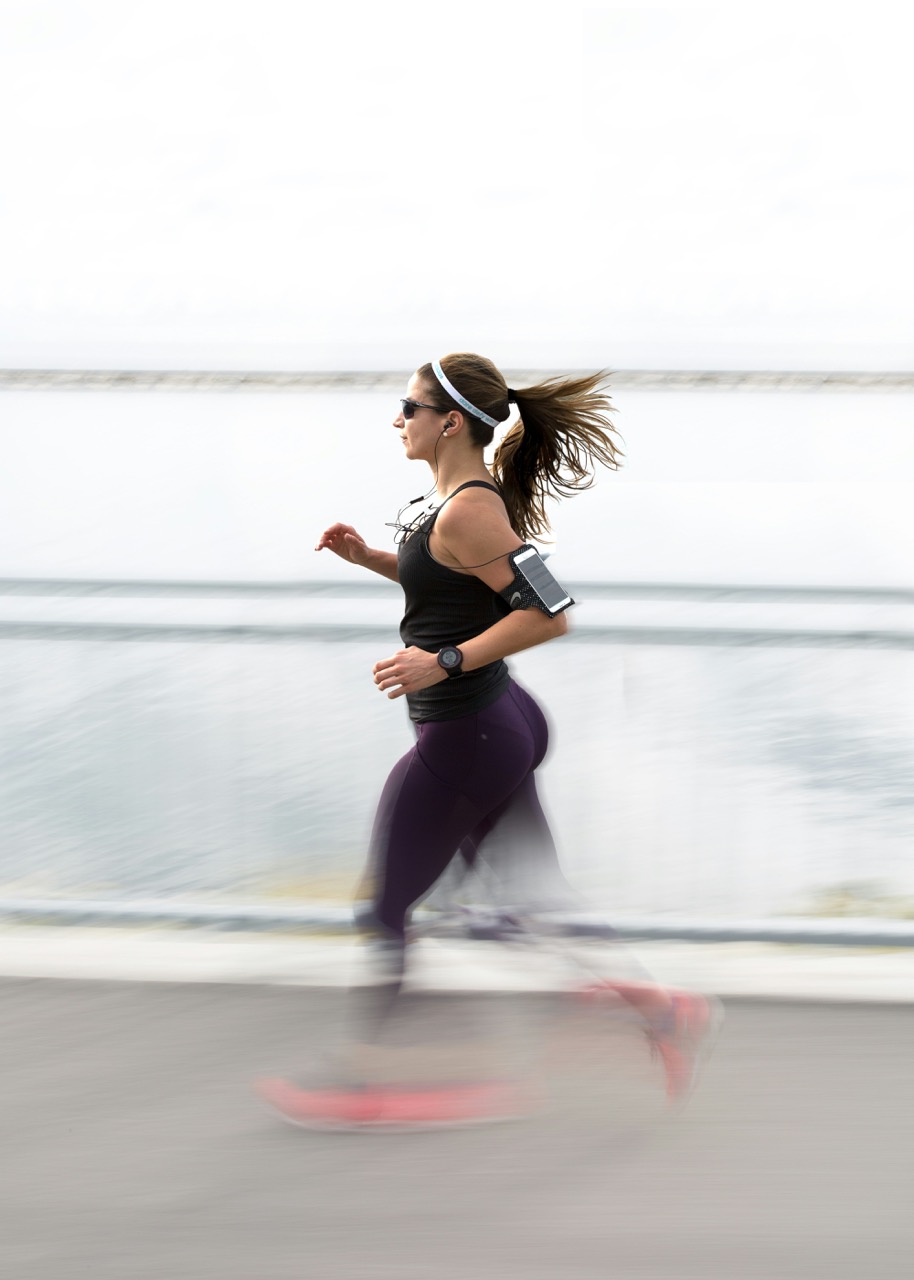 A woman in black sportswear and sunglasses runs near a body of water, her hair flowing behind her, with a motion blur effect.